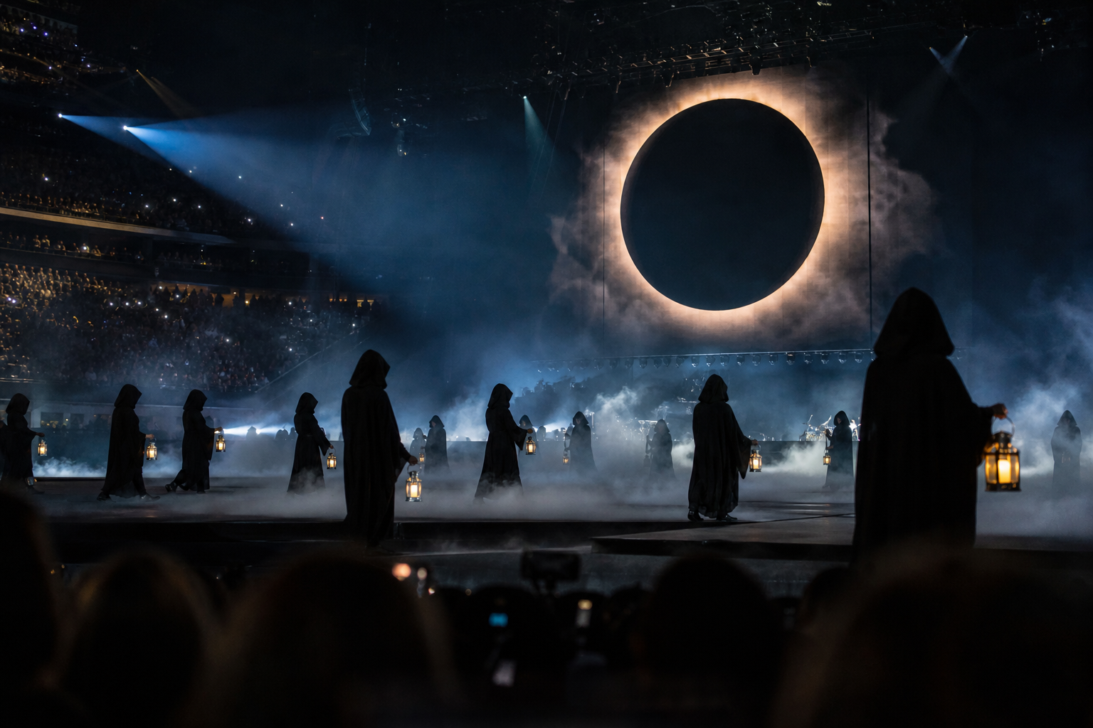 Hooded figures hold dim lanterns on a foggy, dark concert stage under dramatic beam lighting in a stadium arena.
