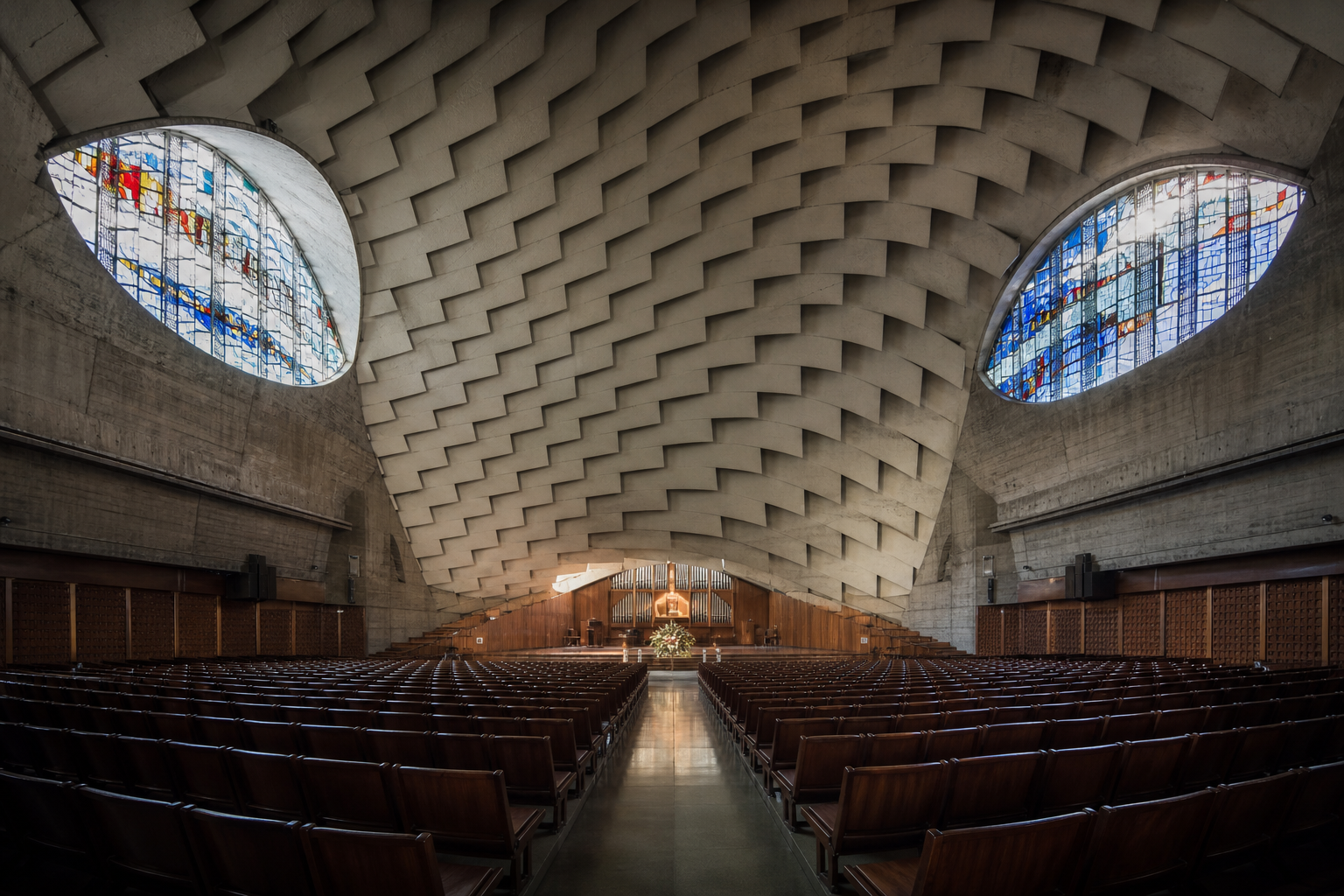 Interior photo of a vast modernist concrete hall with sweeping ribbed vaulted ceilings, oval side windows, and empty seating rows.